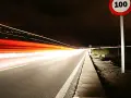 Nocna droga z długimi smugami świateł samochodowych. Znak ograniczenia prędkości 100 km/h przypomina o przepisach, jak ograniczenia prędkości w Holandii.