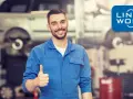 Uśmiechnięty mechanik polski w Holandii daje kciuk w górę. W tle warsztat samochodowy z podniesionym autem.