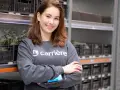 Young woman in a grey sweatshirt with "carrière" logo, holding a scanner, in a warehouse with shelves of black crates.