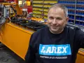 Uśmiechnięty pracownik w bluzie z logo Larex Deventer stoi obok pomarańczowej maszyny w warsztacie.