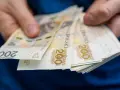 Dłonie trzymają stos banknotów 200 zł. Zastanawiasz się, ile wynosi zasiłek dla bezrobotnych?