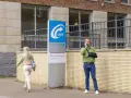 Mężczyzna z ręką na temblaku rozmawia przez telefon przed budynkiem UWV. Czy UWV sprawdza konto bankowe? Kobieta idzie obok.