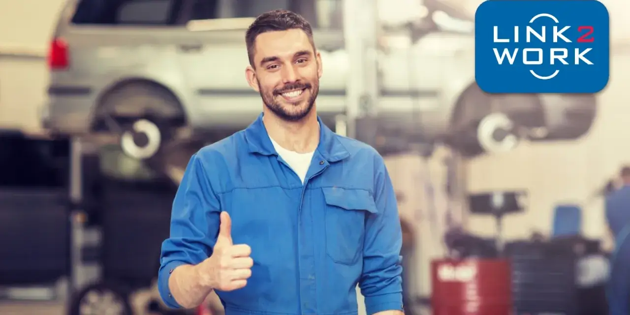 Uśmiechnięty mechanik polski w Holandii daje kciuk w górę. W tle warsztat samochodowy z podniesionym autem.