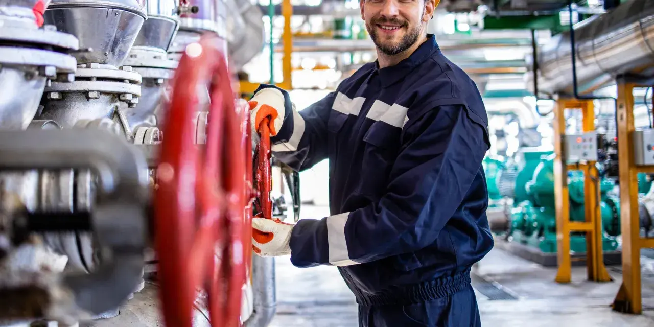 Vrolijke operator koparki holandia, met gele helm, draait aan rode kraan in industriële omgeving.