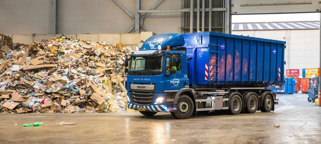 Niebieska ciężarówka z kontenerem firmy Renewi przy ogromnej hałdzie makulatury. Praca w Rotterdamie bezpośrednio związana z recyklingiem.