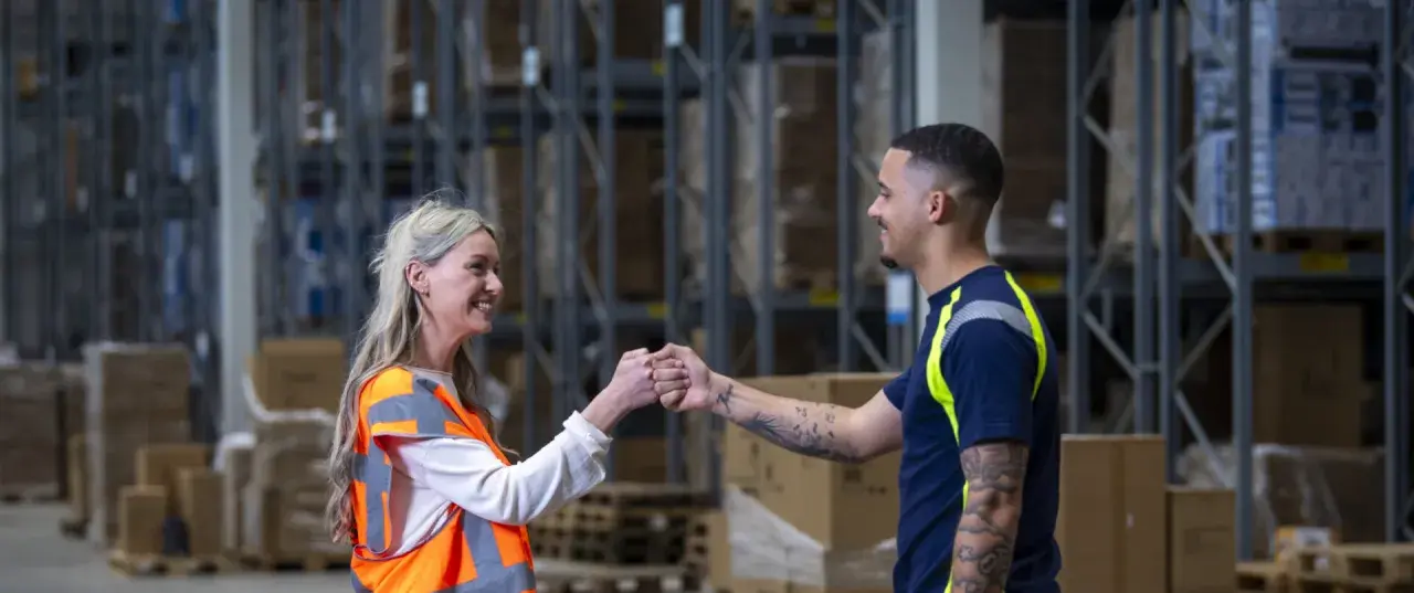 Twee collega's geven elkaar een vuistje in een magazijn. Ze werken in de logistiek en vinden hier hun logistic force oferty pracy.