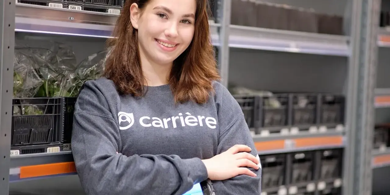 Uśmiechnięta dziewczyna w bluzie z logo Carrière, pracująca w magazynie. Sprawdza zamówienia na skanerze. Szuka kariere oferty pracy.