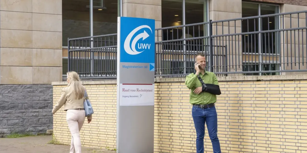 Mężczyzna z ręką na temblaku rozmawia przez telefon przed budynkiem UWV. Czy UWV sprawdza konto bankowe? Kobieta idzie obok.