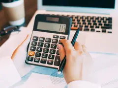 Ręce liczą na kalkulatorze, gdy nie trzeba wykazywać dochodów z zagranicy. Obok laptop, okulary i kubek kawy.