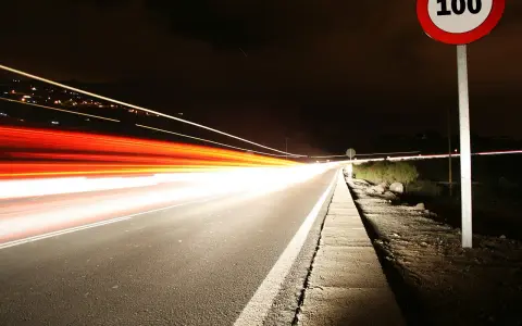 Nocna droga z długimi smugami świateł samochodowych. Znak ograniczenia prędkości 100 km/h przypomina o przepisach, jak ograniczenia prędkości w Holandii.