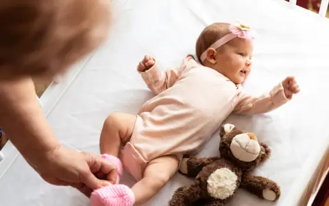 Małe dziecko urodzone w Holandii, w różowym body i opasce z kokardką, leży na białej pościeli obok pluszowego misia.