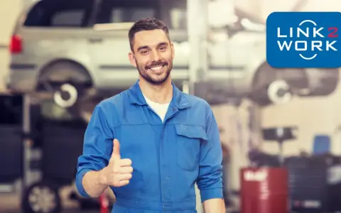 Uśmiechnięty mechanik polski w Holandii daje kciuk w górę. W tle warsztat samochodowy z podniesionym autem.