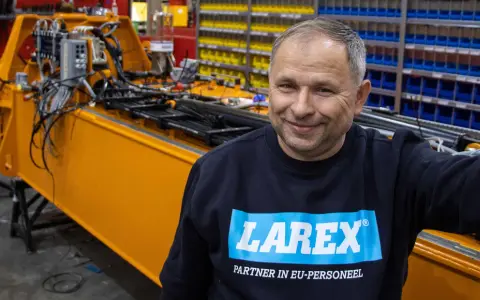 Uśmiechnięty pracownik w bluzie z logo Larex Deventer stoi obok pomarańczowej maszyny w warsztacie.