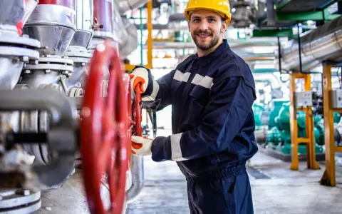 Vrolijke operator koparki holandia, met gele helm, draait aan rode kraan in industriële omgeving.