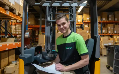 Uśmiechnięty pracownik magazynu, który rozpoczął swoją **dzienną zmianę**, siedzi na wózku widłowym wśród regałów z paczkami.