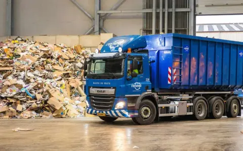 Niebieska ciężarówka z kontenerem firmy Renewi przy ogromnej hałdzie makulatury. Praca w Rotterdamie bezpośrednio związana z recyklingiem.