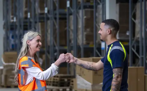 Twee collega's geven elkaar een vuistje in een magazijn. Ze werken in de logistiek en vinden hier hun logistic force oferty pracy.
