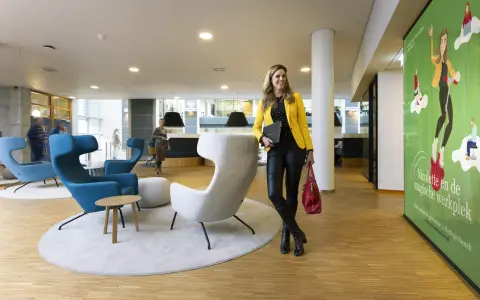 Kobieta w żółtej marynarce i czarnych spodniach stoi w nowoczesnym biurze. Obok niej grafika zachęcająca do pracy w 's-Hertogenbosch.