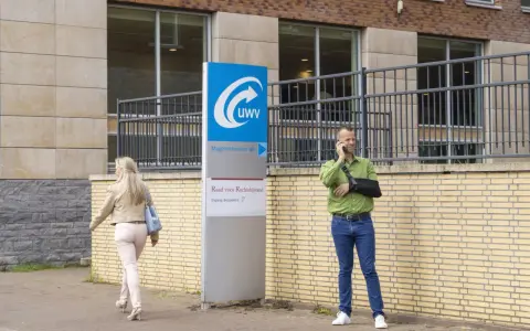 Mężczyzna z ręką na temblaku rozmawia przez telefon przed budynkiem UWV. Czy UWV sprawdza konto bankowe? Kobieta idzie obok.