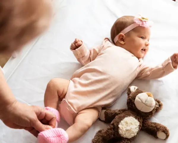 Małe dziecko urodzone w Holandii, w różowym body i opasce z kokardką, leży na białej pościeli obok pluszowego misia.