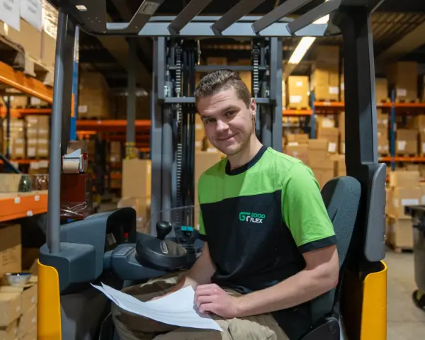 Uśmiechnięty pracownik magazynu, który rozpoczął swoją **dzienną zmianę**, siedzi na wózku widłowym wśród regałów z paczkami.