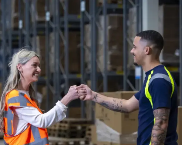 Twee collega's geven elkaar een vuistje in een magazijn. Ze werken in de logistiek en vinden hier hun logistic force oferty pracy.
