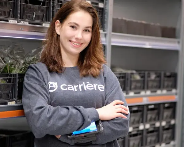 Uśmiechnięta dziewczyna w bluzie z logo Carrière, pracująca w magazynie. Sprawdza zamówienia na skanerze. Szuka kariere oferty pracy.