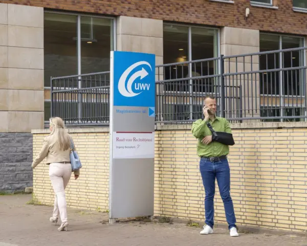 Mężczyzna z ręką na temblaku rozmawia przez telefon przed budynkiem UWV. Czy UWV sprawdza konto bankowe? Kobieta idzie obok.