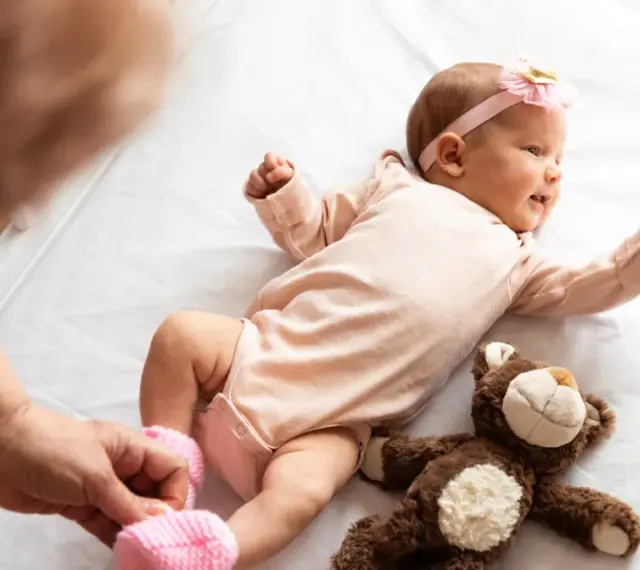 Małe dziecko urodzone w Holandii, w różowym body i opasce z kokardką, leży na białej pościeli obok pluszowego misia.