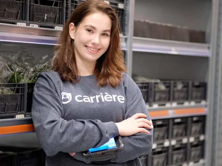 Uśmiechnięta dziewczyna w bluzie z logo Carrière, pracująca w magazynie. Sprawdza zamówienia na skanerze. Szuka kariere oferty pracy.