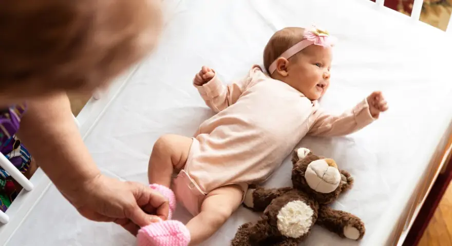 Małe dziecko urodzone w Holandii, w różowym body i opasce z kokardką, leży na białej pościeli obok pluszowego misia.
