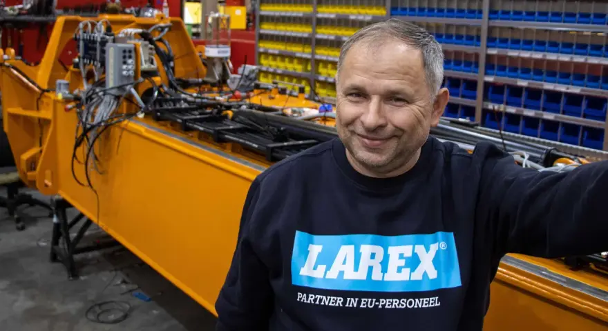 Uśmiechnięty pracownik w bluzie z logo Larex Deventer stoi obok pomarańczowej maszyny w warsztacie.