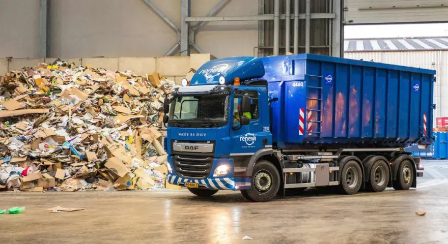 Niebieska ciężarówka z kontenerem firmy Renewi przy ogromnej hałdzie makulatury. Praca w Rotterdamie bezpośrednio związana z recyklingiem.