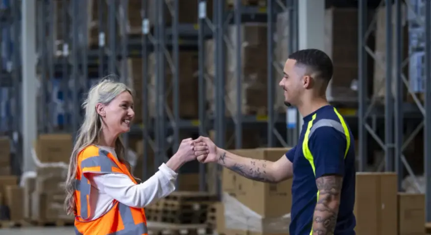 Twee collega's geven elkaar een vuistje in een magazijn. Ze werken in de logistiek en vinden hier hun logistic force oferty pracy.
