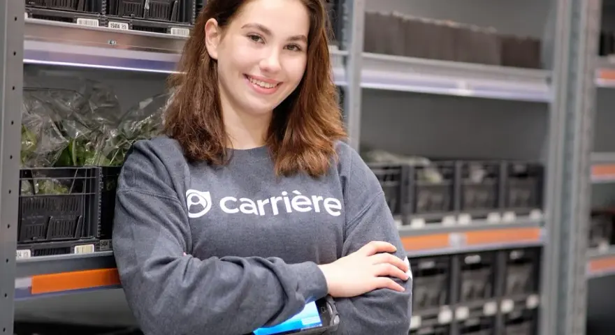 Uśmiechnięta dziewczyna w bluzie z logo Carrière, pracująca w magazynie. Sprawdza zamówienia na skanerze. Szuka kariere oferty pracy.