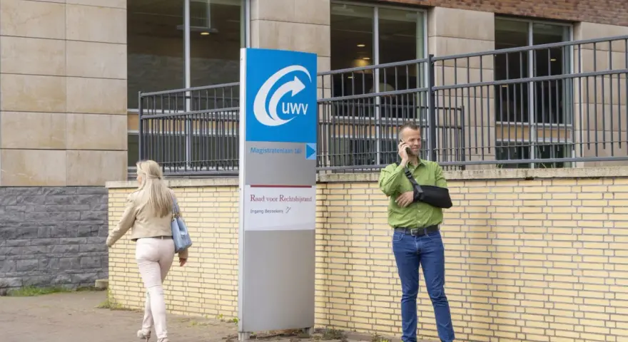 Mężczyzna z ręką na temblaku rozmawia przez telefon przed budynkiem UWV. Czy UWV sprawdza konto bankowe? Kobieta idzie obok.