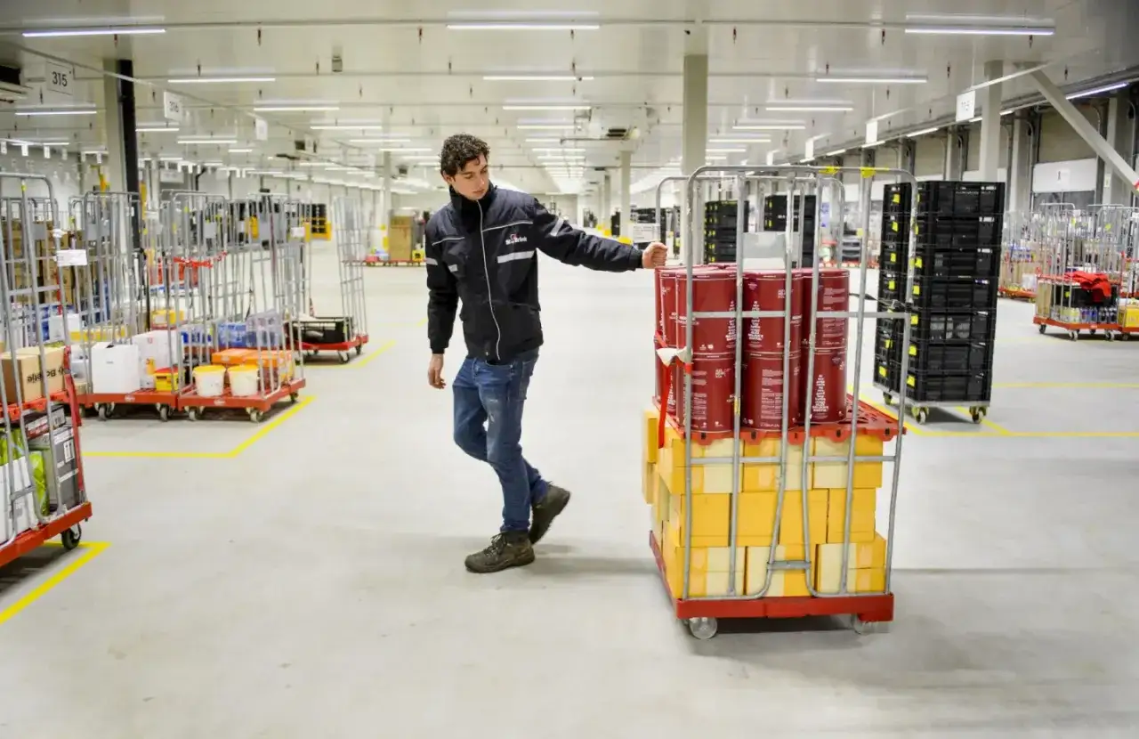 Pracownik w magazynie GXO Nieuwegein przesuwa wózek z paczkami i czerwonymi pojemnikami.