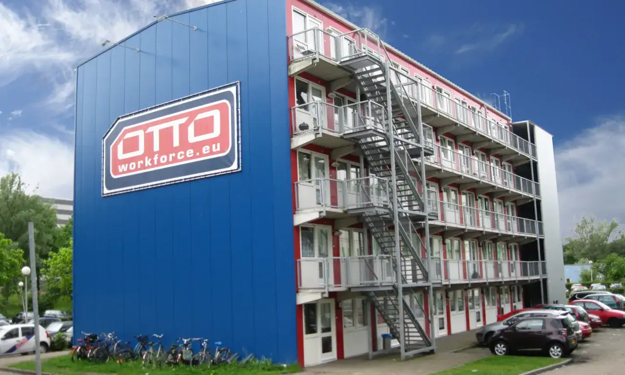 Budynek z niebieskim panelem i logo OTTO Workforce.eu, oferujący eb personeelsdiensten. Wiele rowerów i samochodów na parkingu.