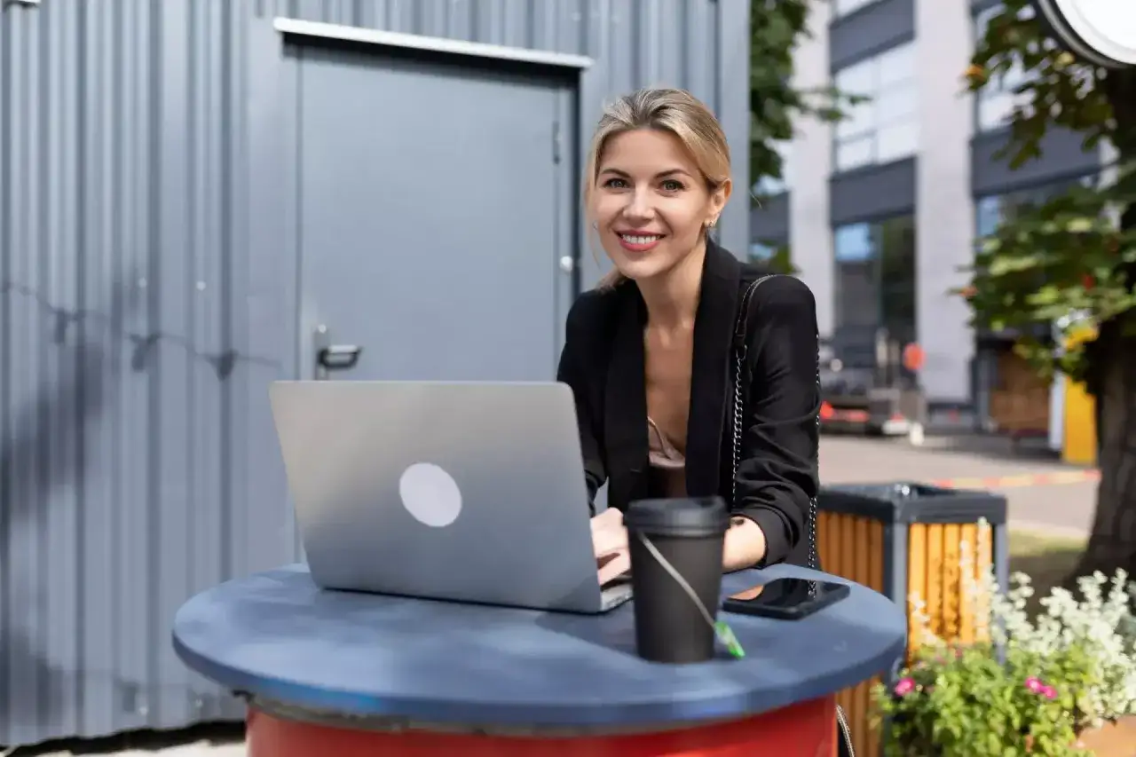 Uśmiechnięta kobieta pracuje na laptopie na zewnątrz. Analizuje zzp holandia koszty, planując swoje finanse.