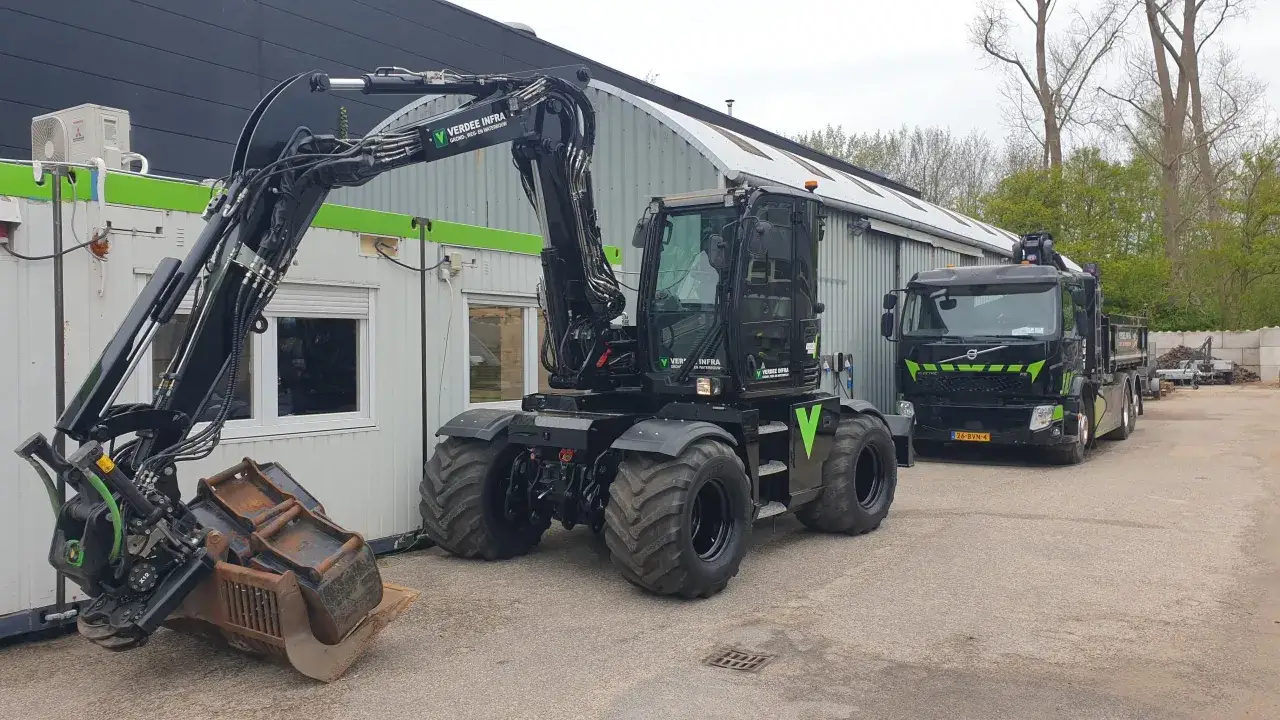 Zwarte mobiele graafmachine met grote banden, klaar voor werk. Een vrachtwagen staat ernaast. Een operator koparki hollandse klus.