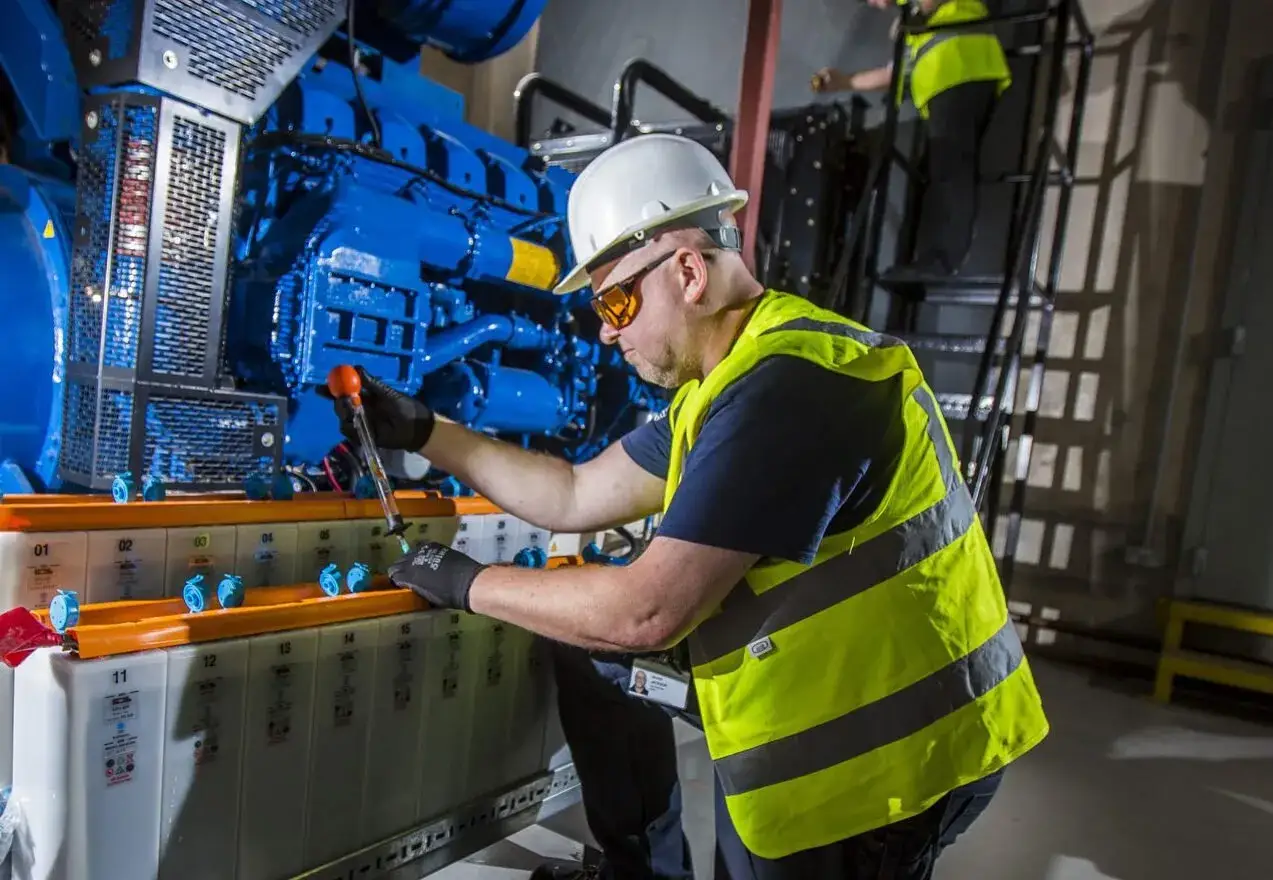 Technicus controleert batterijen bij een grote generator. Een collega werkt op een ladder. Goede kansen bij Larex!