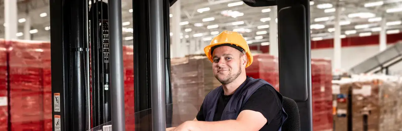 Uśmiechnięty pracownik w ż&oacute;łtym kasku siedzi w w&oacute;zku widłowym, gotowy do pracy w magazynie. To jego praca w Limburgii.
