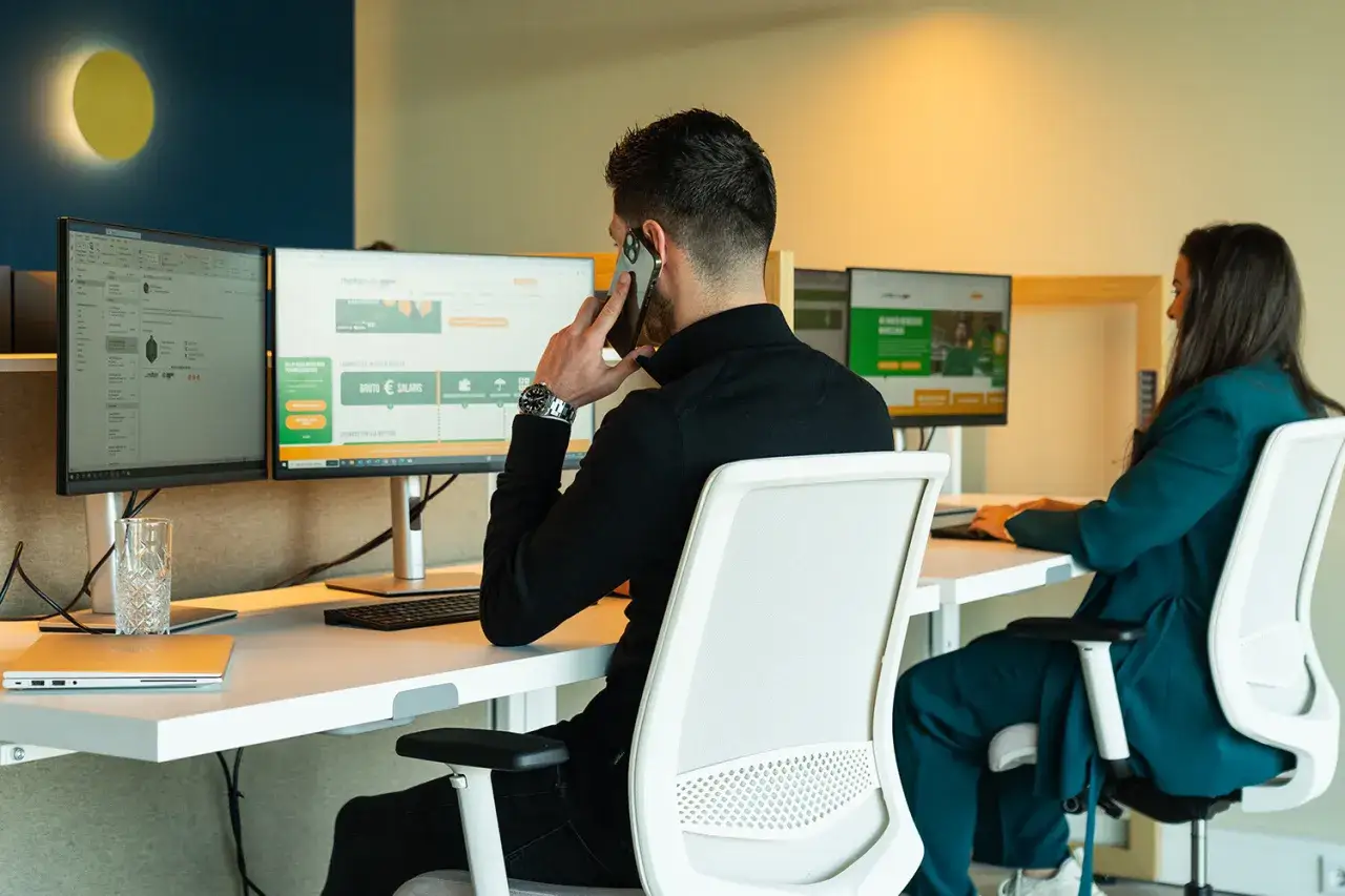 Mężczyzna rozmawia przez telefon, pracując przy komputerze. Kobieta siedzi obok, również przy biurku. Planują urlop na żądanie w Holandii.
