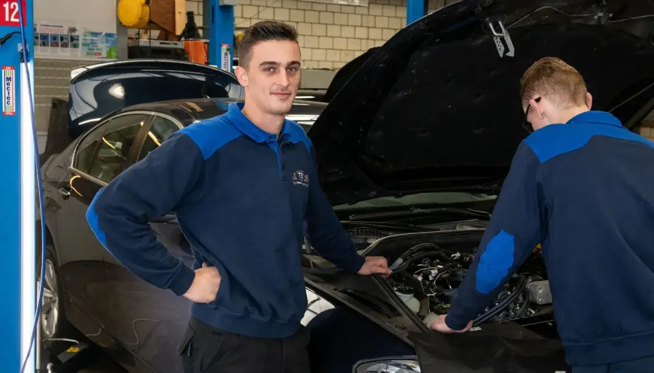 Młody mechanik polski w Holandii, z uśmiechem przygląda się pracy kolegi nad silnikiem samochodu.