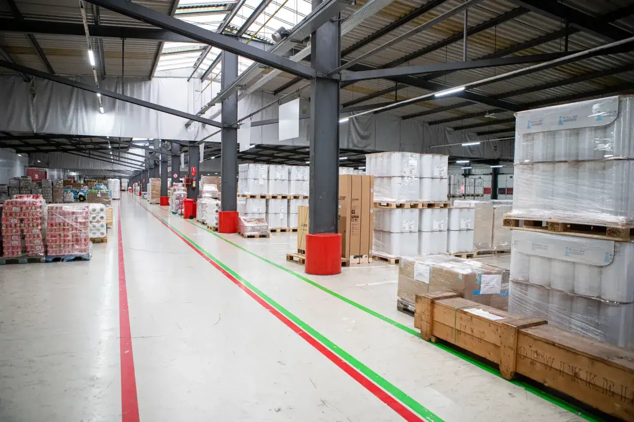 Wielki magazyn w Dordrecht, pełen palet z towarami. Praca wre, linie na podłodze wyznaczają ścieżki.