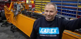 Uśmiechnięty pracownik w bluzie z logo Larex Deventer stoi obok pomarańczowej maszyny w warsztacie.