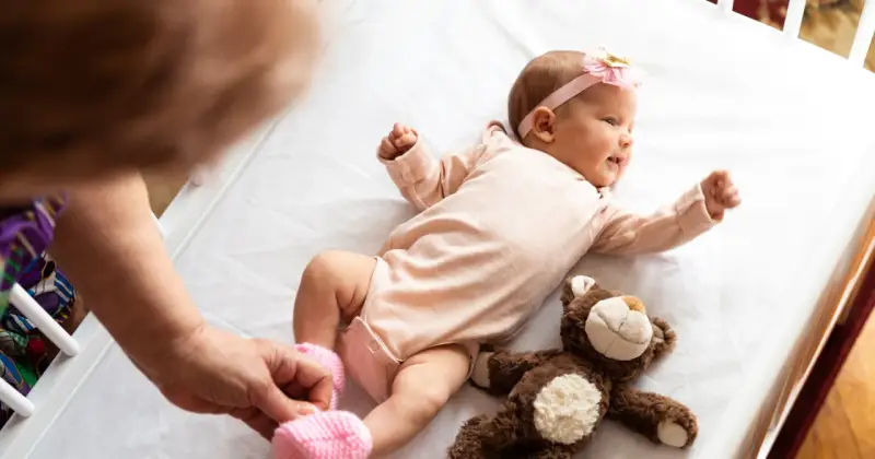 Małe dziecko urodzone w Holandii, w różowym body i opasce z kokardką, leży na białej pościeli obok pluszowego misia.