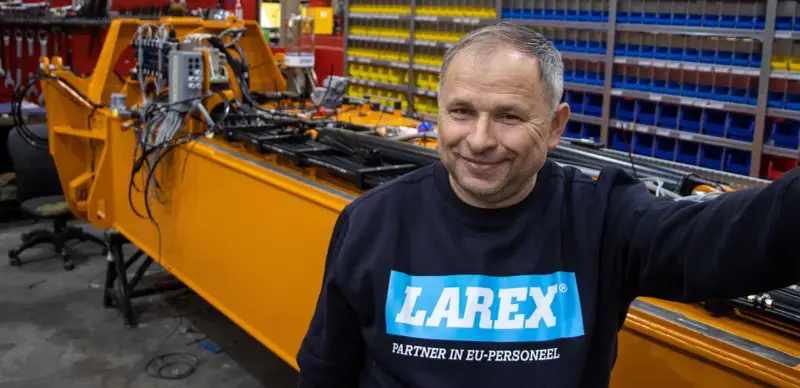 Uśmiechnięty pracownik w bluzie z logo Larex Deventer stoi obok pomarańczowej maszyny w warsztacie.